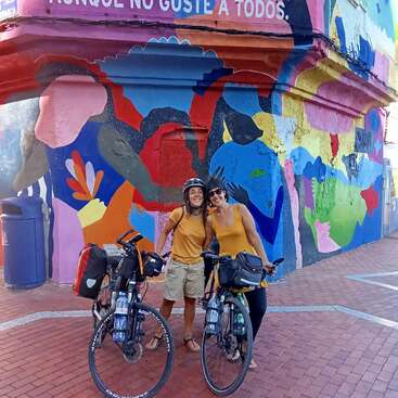 Two people with bicycles smile in front of a vibrant, colorful mural. Both wear yellow tops. The street scene feels cheerful, artistic, and full of energy.