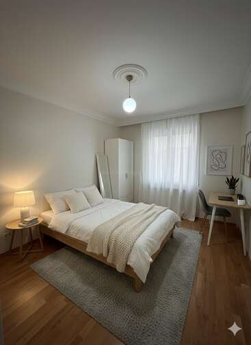 This cozy bedroom features a wooden bed with white bedding, a bedside lamp, desk, chair, wardrobe, standing mirror, abstract art, and soft natural lighting.