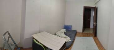 This image shows a simple, sparsely furnished room with a mattress on the floor, pillow, blanket, desk, chair, step ladder, and a rug. Door open.