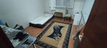 This is a small, simple room with a mattress on the floor, chair, desk, golden table, patterned rug, white curtain, and hardwood floor.