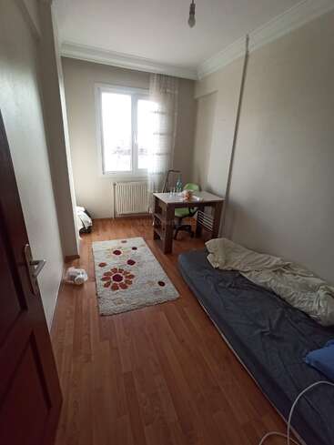 A small, simple room with a mattress on the floor, a desk and chair by the window, a rug, and minimal decorations. Sunlight enters.