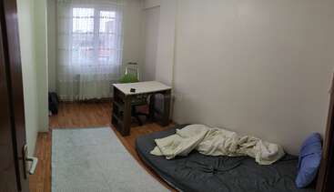 A simple, minimally furnished room with a mattress on the floor, a blue pillow, and a messy blanket. A desk and chair sit by the window.