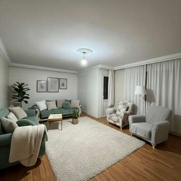 Ce salon confortable comprend deux canapés verts, deux fauteuils, un tapis blanc, du parquet, des plantes en pot, des œuvres d'art murales encadrées et des rideaux blancs allant du sol au plafond.