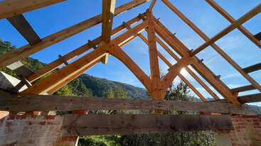 A partially constructed wooden roof frame sits atop brick walls, open to the sky. Sunlight shines through rafters, with green hills and blue skies in the background.