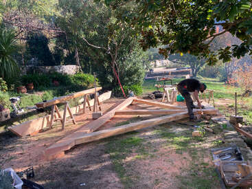 A person works outdoors on a wooden structure, surrounded by trees and tools. Several sawhorses support lumber. The setting is sunny, leafy, and tranquil.