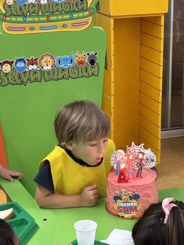 A young boy in a yellow vest blows out a candle on a pink Fireman Sam-themed birthday cake with Spider-Man decorations at a colorful party.