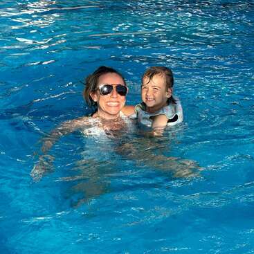 Eine Frau mit Sonnenbrille und ein lächelndes Kind in einer Schwimmweste schwimmen zusammen in einem hellblauen Pool und wirken glücklich und entspannt.