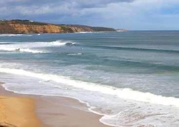A imagem retrata uma cena serena de praia com ondas suaves rolando na costa, tendo como pano de fundo um penhasco e um céu nublado.