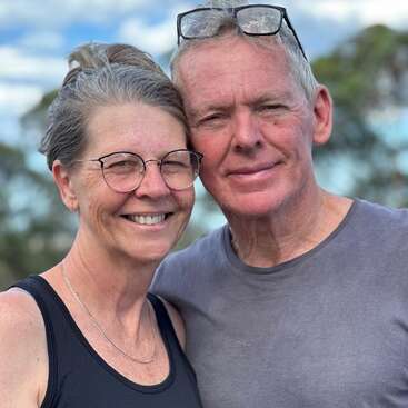 A smiling couple stands close together outdoors. The woman wears glasses and a black top; the man has glasses on his head and a gray shirt.
