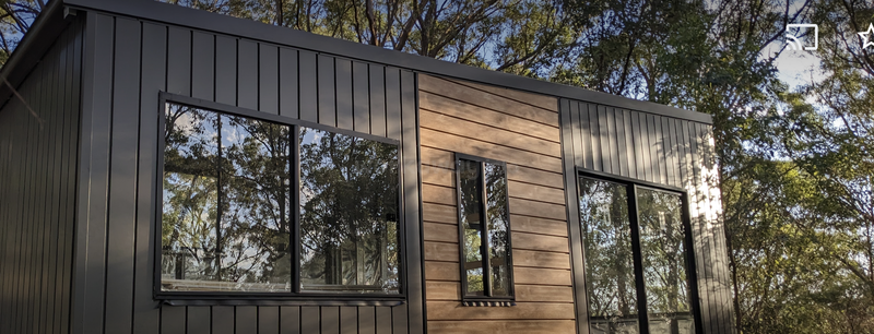 This image shows a modern tiny house with large glass windows, black vertical siding, and a wood accent panel, surrounded by tall, leafy trees under sunlight.
