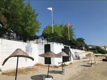 Ein Sandstrand mit mehreren Strohsonnenschirmen, ein weißes Gebäude mit schwarzen Türen, Grünpflanzen darüber und Fahnen, die an einem sonnigen Tag am blauen Himmel wehen.