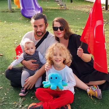 A family of four sits on grass at a playground. The mother holds a Turkish flag, the father holds a baby, and the older child smiles with a toy octopus.