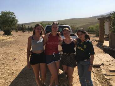 Cuatro mujeres jóvenes están muy juntas, sonrientes, delante de una camioneta blanca en un día soleado en un paisaje rural y árido con colinas y árboles.