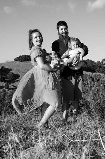 Eine lächelnde vierköpfige Familie steht barfuß auf einer Wiese. Die Mutter hält ein Baby, der Vater ein Kleinkind. Es ist ein fröhlicher, windiger Tag.