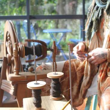 L'image représente une femme en train de filer du fil sur un rouet en bois, entourée de deux bobines de fil et d'une table sur laquelle se trouve un crayon.