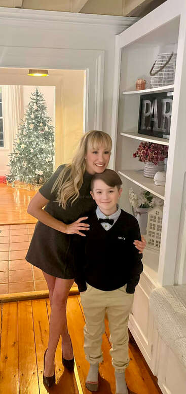 Una mujer con un vestido negro y un niño con jersey y pajarita posan sonrientes, con un árbol de Navidad decorado iluminado al fondo.