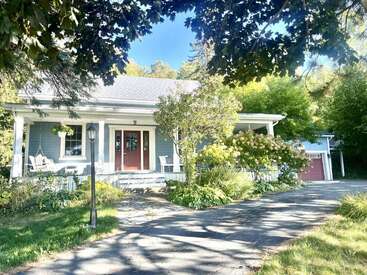 A charming blue house with a welcoming front porch, red double doors, lush landscaping, swing seat, and a driveway leading to a garage, surrounded by trees.