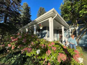 Une femme se tient dans un jardin luxuriant, près d'une charmante maison bleue ornée de colonnes blanches, d'arbustes en fleurs et de paniers suspendus, sous un ciel bleu limpide.