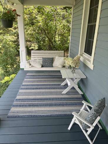 Une véranda chaleureuse est agrémentée d'une balancelle blanche avec des coussins, d'un tapis à rayures bleues, d'une petite table ornée de fleurs, d'une chaise, d'une plante suspendue et d'une végétation luxuriante à l'extérieur.