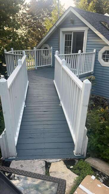 Une rampe en bois bleu, bordée de balustrades blanches, mène à un chalet au toit à pignon et aux portes vitrées, entouré de verdure et d'un escalier en pierre naturelle.