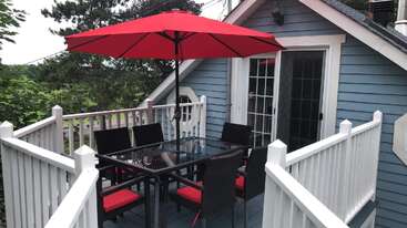Cette image montre un balcon extérieur chaleureux avec une table en verre, six chaises noires dotées de coussins rouges et un grand parasol rouge, adossé à une maison bleue.