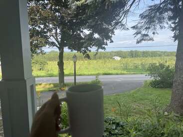 Sur un porche, une main tient une tasse blanche, donnant sur une route tranquille, un champ verdoyant, des arbres et un paysage rural paisible et ensoleillé sous un ciel bleu.