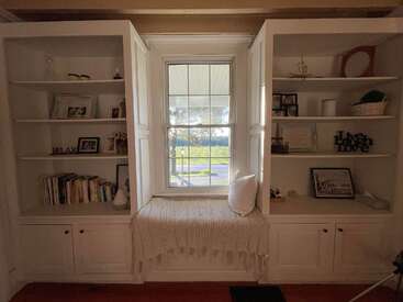 Un coin fenêtre douillet est niché entre deux bibliothèques encastrées blanches regorgeant de livres, de photos encadrées et d'objets décoratifs, baigné par la douce lumière du soleil qui y pénètre.