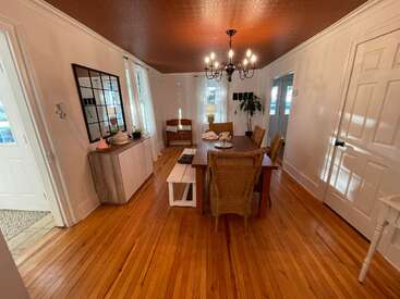 Cette salle à manger est dotée d'un parquet, d'une table rectangulaire, de chaises en osier, d'un lustre, de murs blancs, de grandes fenêtres, d'un buffet et de plantes décoratives qui lui confèrent une touche d'élégance.