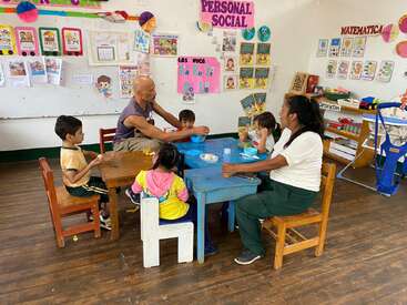 Dans une salle de classe colorée, deux adultes et cinq jeunes enfants sont assis autour de petites tables, mangeant et discutant. Des affiches éducatives et des dessins d'enfants décorent les murs animés.