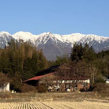 Uma casa rústica fica entre árvores e campos, com montanhas cobertas de neve ao fundo, sob um céu azul límpido, representando um cenário rural tranquilo e a beleza da natureza.