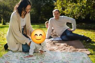 Una familia disfruta de un picnic al aire libre bajo el sol. La madre ayuda a su bebé a gatear sobre una manta, mientras el padre y otro niño descansan al fondo.