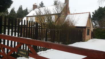 Una pintoresca casa de ladrillo con el tejado cubierto de nieve está rodeada de árboles y una valla de madera oscura. La nieve cubre el jardín y la verja roja.