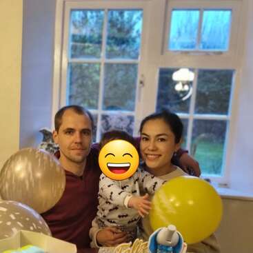 Uma família feliz de três pessoas está sentada junto à janela, segurando balões. O rosto da criança está coberto por um emoji sorridente, criando uma atmosfera alegre.