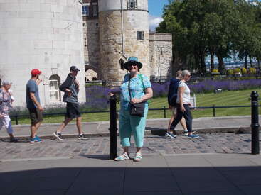 A woman in turquoise stands smiling in front of historic stone towers, while several people walk along a cobblestone path. Lavender flowers bloom nearby. Sunny day.