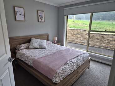 Dieses gemütliche Schlafzimmer verfügt über ein Holzbett mit Bettwäsche mit Blumenmuster, eine zartrosa Decke, ein Kissen, eine Nachttischlampe, Wandbilder und ein großes Fenster mit Blick ins Grüne.