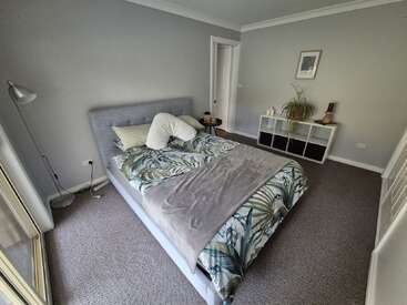 Ein gemütliches Schlafzimmer mit einem grauen Bett, Bettwäsche mit Blattmuster, einem Nachttisch, einer Stehlampe, Regalen, einer grünen Pflanze, Kunstwerken und einem Teppichboden in neutralen Farbtönen.