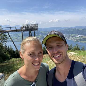 A smiling couple takes a selfie outdoors with a scenic background of hills, a lake, forests, and a wooden observation platform under a bright sunny sky.
