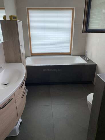 Dieses moderne Badezimmer verfügt über eine elegante Badewanne unter einem großen, schattigen Fenster, einen geschwungenen Waschbeckenschrank, einen Wandspiegel, eine Toilette, großzügige graue Fliesen und eine minimalistische Einrichtung.