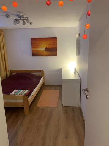 Ein gemütliches Schlafzimmer mit Holzboden, Einzelbett mit kastanienbrauner Decke, Kommode, orangefarbenem Teppich, Sonnenuntergangsgemälde, Lichterketten, moderner Deckenlampe und warmer Raumbeleuchtung.