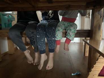 Drei Kinder in Schlafanzügen sitzen auf einer Bank unter einem Holztisch, ihre nackten Füße baumeln und schaffen einen gemütlichen, verspielten und ehrlichen Familienmoment im Haus.