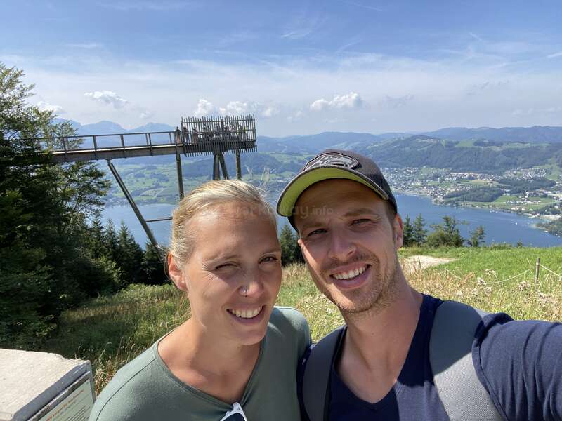 Ein lächelndes Paar macht ein Selfie im Freien vor einem malerischen Hintergrund aus Hügeln, einem See, Wäldern und einer hölzernen Aussichtsplattform unter einem strahlenden Sonnenhimmel.
