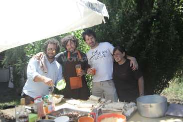 Das Bild zeigt vier Personen, die hinter einem mit Essen und Getränken beladenen Tisch stehen, vor einer Kulisse aus Bäumen und einem weißen Regenschirm.
