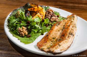Das Bild zeigt einen Teller mit Essen auf einem Holztisch, auf dem ein Salat mit gemischtem Grün, Walnüssen, Rosinen und gegrillten Orangenscheiben liegt, begleitet von einem gegrillten Fischfilet.