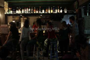 Das Bild zeigt eine schwach beleuchtete Barszene, in der Gäste an der Bar sitzen und ein Barkeeper hinter dem Tresen zu sehen ist.