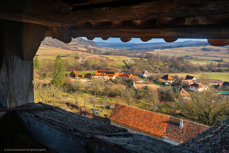 A imagem mostra uma charmosa vila rural cercada por colinas onduladas, casas tradicionais com telhados vermelhos, vegetação exuberante e campos distantes, vista de uma estrutura rústica de madeira.