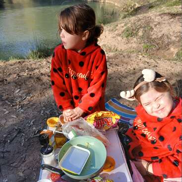 Two young girls wearing red, ladybug-patterned hoodies sit by a riverside picnic table, enjoying snacks and sunlight. One girl squints, the other gazes away.