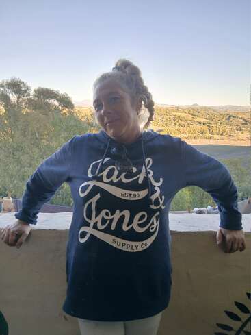 A woman stands on a balcony wearing a blue "Jack & Jones" hoodie. Behind her is a scenic landscape with trees, hills, and a clear sky.