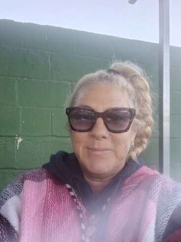 A woman with blonde braided hair and sunglasses smiles at the camera. She wears a pink and white woven poncho and stands in front of a green wall.