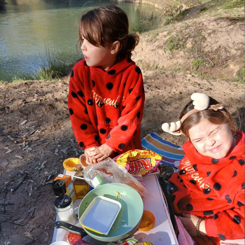 Two young girls wearing red, ladybug-patterned hoodies sit by a riverside picnic table, enjoying snacks and sunlight. One girl squints, the other gazes away.