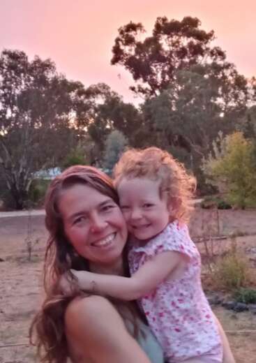 Eine lächelnde Frau hält ein glückliches junges Mädchen im Freien. Der Himmel leuchtet bei Sonnenuntergang rosa, und hohe Bäume rahmen den Hintergrund ein und schaffen eine warme, liebevolle Szene.